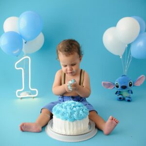 A cute baby enjoying a vibrant blue birthday cake with balloons and toys.