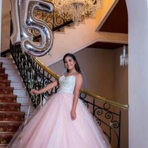Young woman in a gown on stairs with '15' balloons, marking her quinceañera celebration.